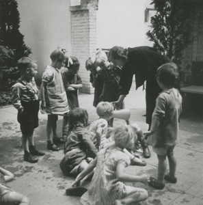 876960 Afbeelding van een groep kleuters rond een Zuster Augustines in haar functie als 'kleuterjuf', die water schenkt ...